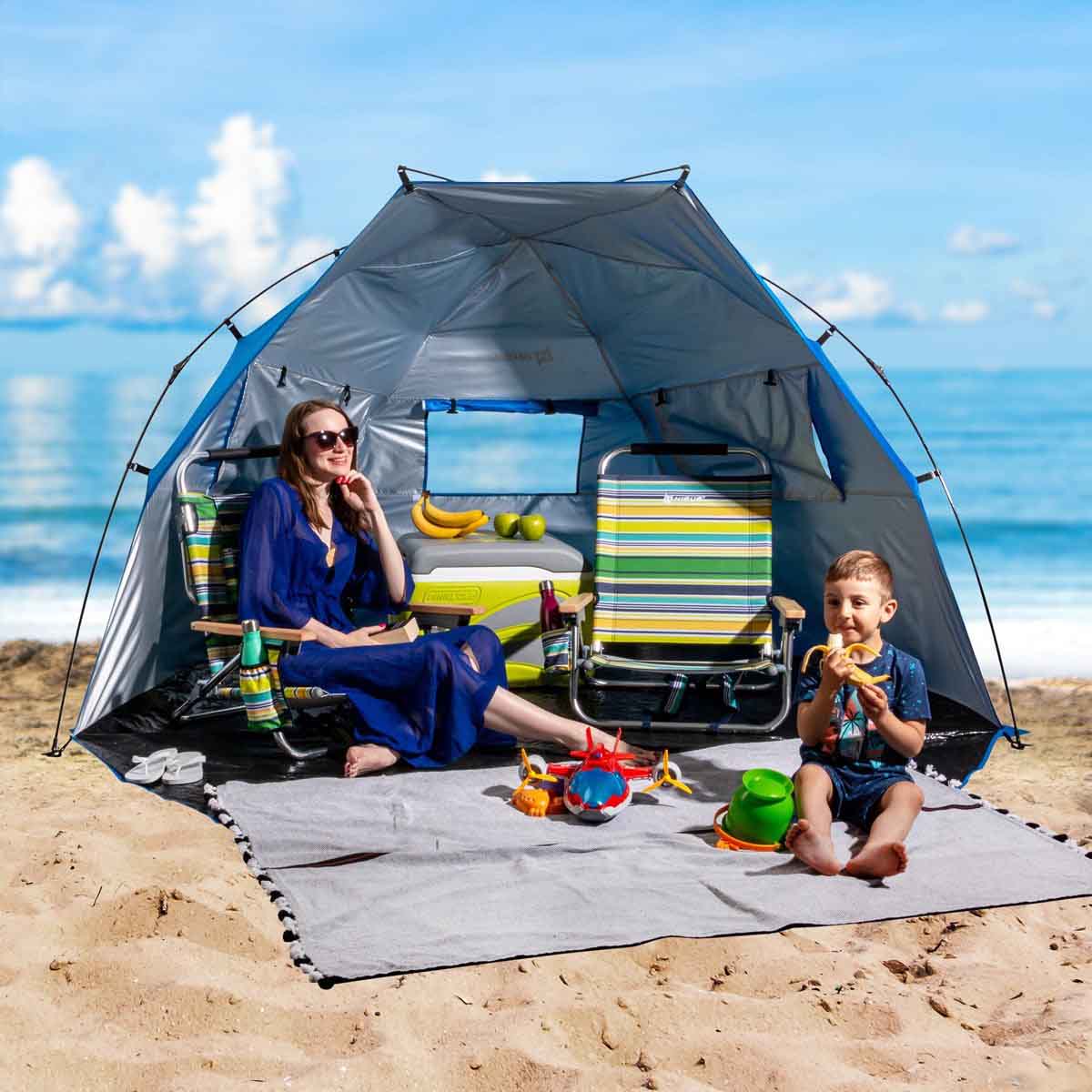 A woman is sitting in the 4 Person Large Easy Up Beach Tent Sun Shade Shelter UPF 50+, a child sitting sitting by eating a banana